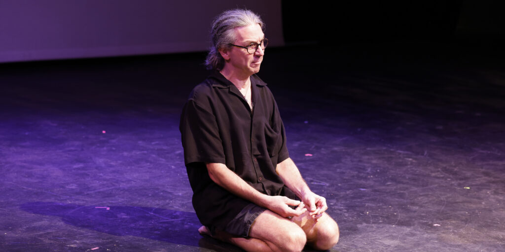 Adam Griebel, pictured here in mid-performance, sitting on his knees as he stares out at the audience.